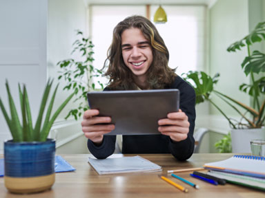 Lachender Abiturient sitzt mit Tablet am Schreibtisch und scheint zu lernen.