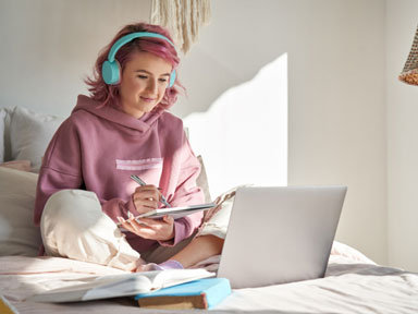 Schülerin mit pinken Haaren und Kopfhörern mit Laptop und Lernmaterialien auf dem Bett